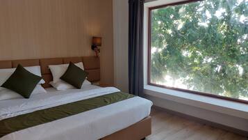 Ein modernes Hotelzimmer mit einem großen Bett, einem hölzernen Kopfteil, einem Nachttisch mit Lampe und einem großen Fenster mit Blick auf Bäume.