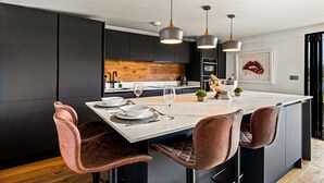 A modern kitchen with a central island, pendant lighting, and a view of the dining area. - River cabin & Mills Cottage (Henley-On-Thames)
