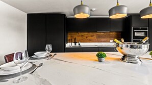 A modern kitchen with black cabinets, a wooden backsplash, and a marble countertop. There are two pendant lights, a silver bowl with champagne, and a small potted plant. - River cabin & Mills Cottage (Henley-On-Thames)