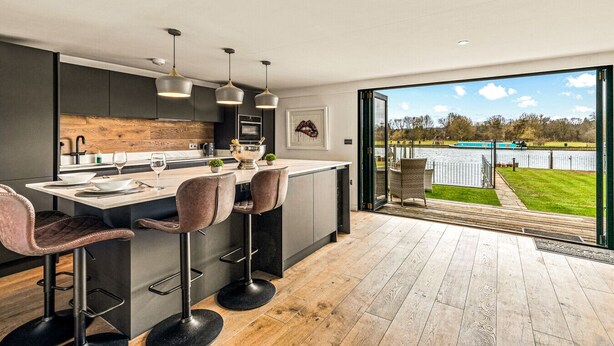 A modern kitchen with a large island, bar stools, pendant lighting, and a view of a lake through an open door. - River cabin & Mills Cottage (Henley-On-Thames)
