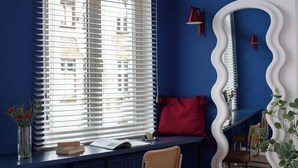 Ein Zimmer mit Fenster und Jalousien, einem roten Kissen, einem Spiegel mit geschwungener Rahmung, einem Stuhl und einem Schreibtisch mit Buch und Blumenvase.