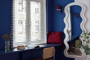 Una habitación con ventana con persianas, almohada roja, espejo con marco ondulado, una silla y un escritorio con un libro y un florero.