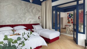 Ein Hotelzimmer mit roter Akzentwand, einem großen Bett, einem kleineren Bett, einem Essbereich mit Tisch und Stühlen sowie Blick durch eine Glastür.