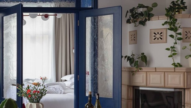 Ein Zimmer mit einem Bett, einem Esstisch mit Früchten und Flaschen, einem Kamin und Blick durch eine türförmige Öffnung mit blauem Rahmen.