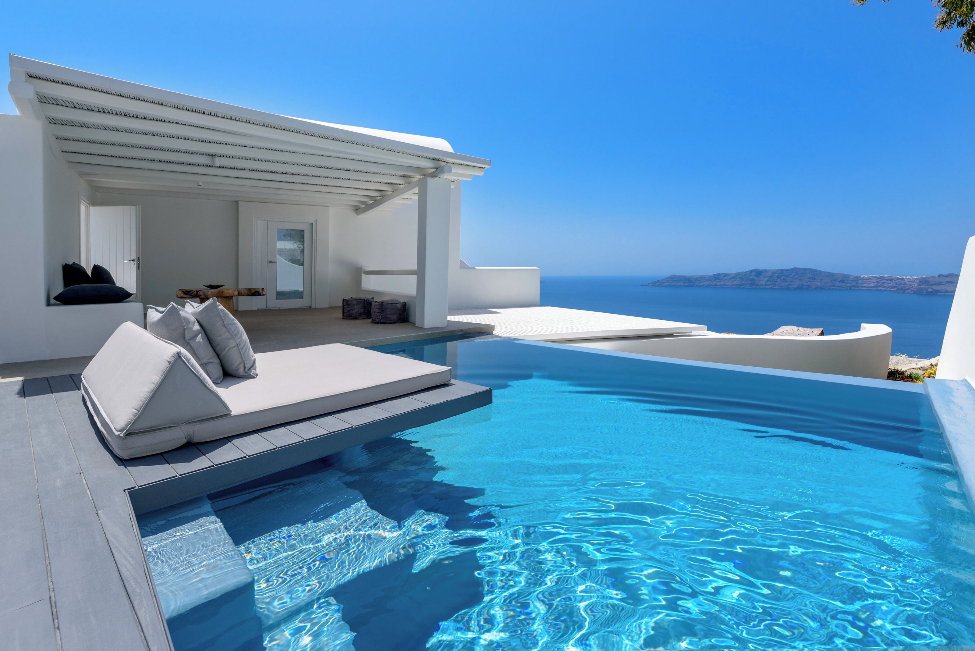A modern infinity pool with a clear view of the sea, a white outdoor seating area with cushions, and a minimalist architectural design.
