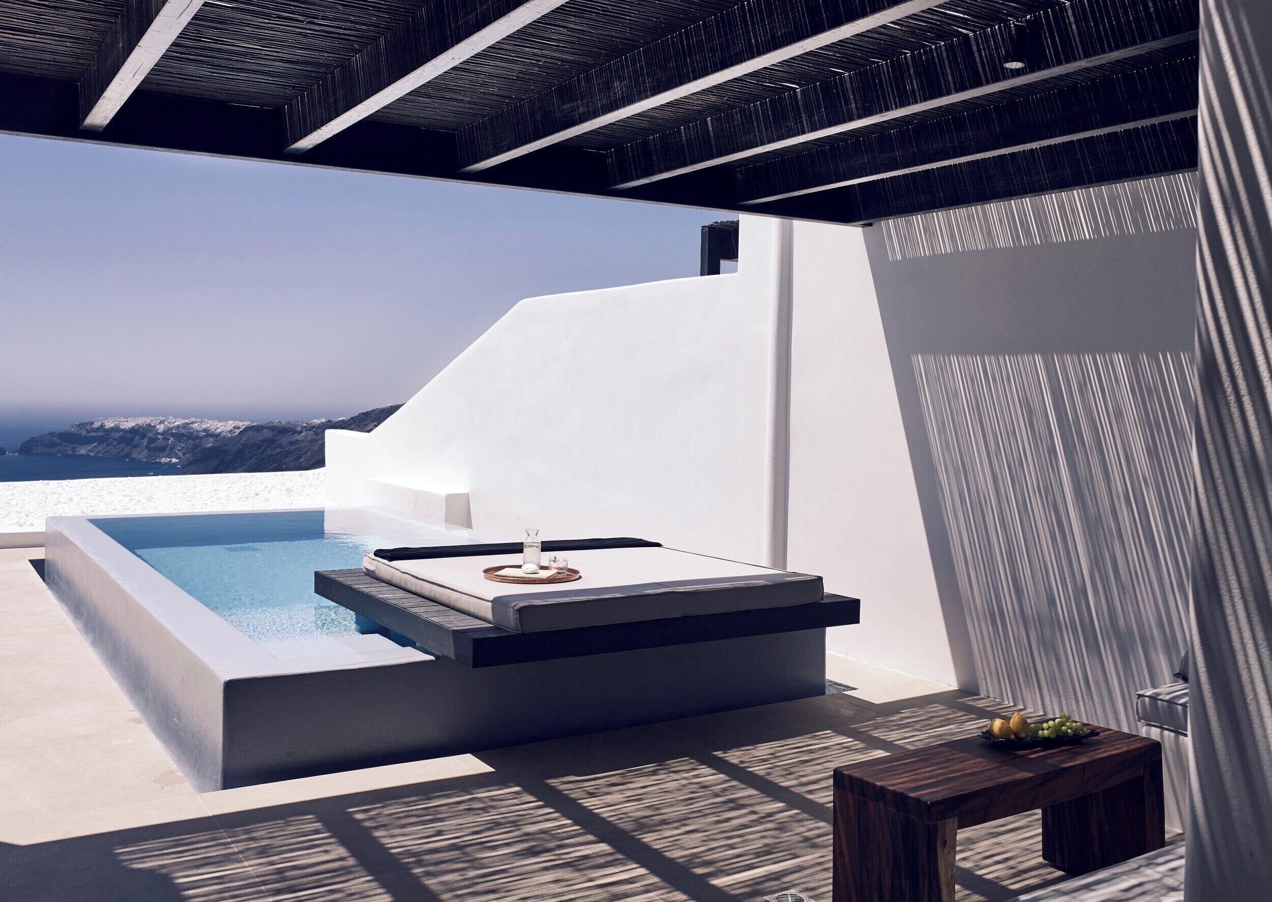 Un espace de détente extérieur moderne, doté d’une piscine, d’un coin salon avec une table basse et d’une petite table sur laquelle sont disposés des fruits.