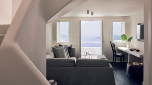 Ein gemütlicher Wohnraum mit einer Couch, einem kleinen Tisch und einem Sessel. Zwei Fenster bieten Blick auf das Meer.