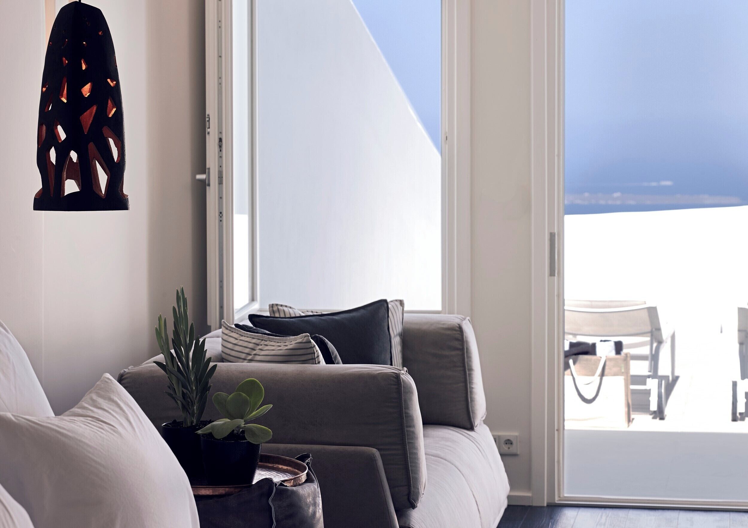 Un salon moderne avec un canapé, des coussins, une plante en pot et une vue sur la mer par une grande fenêtre.