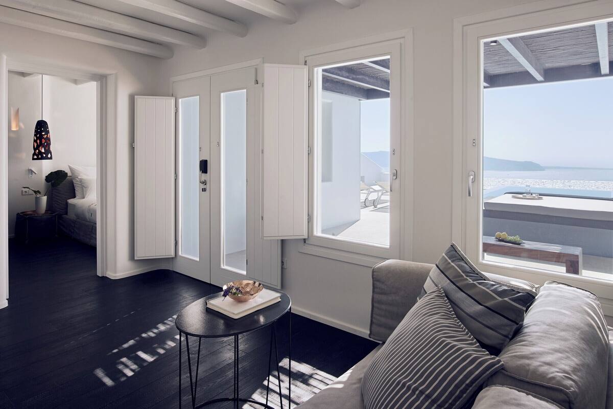 A modern living room with a dark wooden floor, a sofa with striped cushions, a small round table with a bowl and book, and large windows offering a view of the sea.