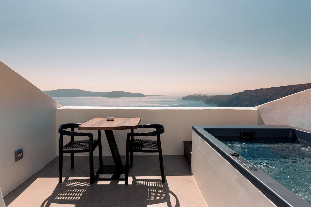 A balcony with a table and chairs, a hot tub, and a view of the sea and hills.