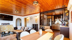 A modern living room with a wooden ceiling, a dining area with a table and chairs, a kitchen, and a TV mounted on the wall. - Seaview Nature Retreat - Filiatra s Cozy Hideaway (Trifylia)