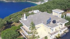 A two-story building with a flat roof, solar panels, and a water tank. It is surrounded by greenery and overlooks a coastal area with a beach and hills. - Ionian View Haven - Parga Paradise (Parga)