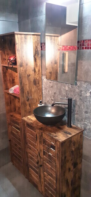 En sminkbord av trä med en svart tvättställ och blandare, en spegel ovanför och en hylla med handdukar.
