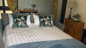 A neatly made bed with a striped bedspread and two floral pillows, a wooden nightstand with a lamp and a vase of flowers, and a view of a window with curtains.