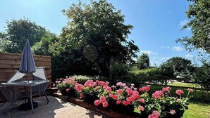 A patio with a table and chairs, surrounded by a flower bed with pink flowers, and a wooden fence in the background.