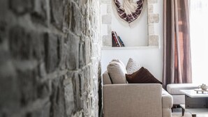 A cozy living room with a stone wall, a sofa with cushions, a small table, and a bookshelf with books and decorative items. - Villa Mira (Kamilari)