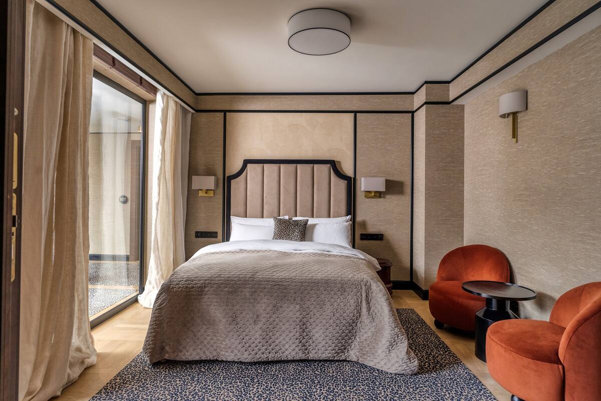 A hotel room with a large bed, a glass door, two side tables, and two orange chairs.