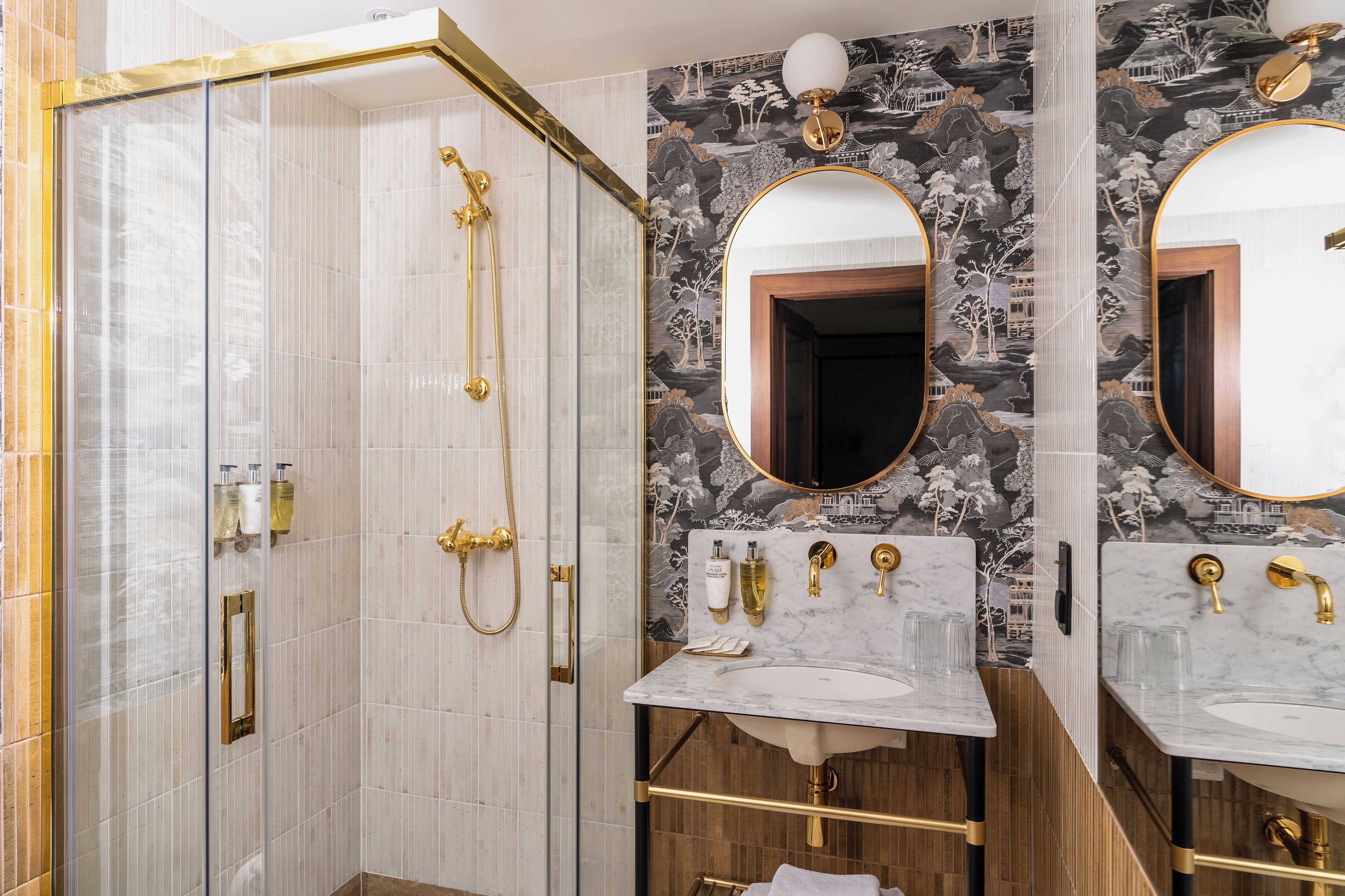 A bathroom with a walk-in shower, two sinks with gold fixtures, and a decorative mirror.