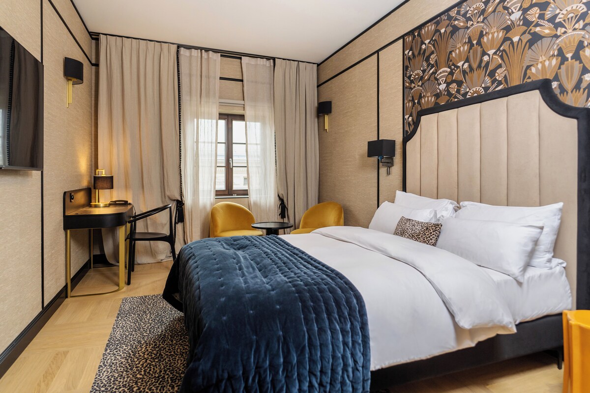 A hotel room with a bed, a desk, two chairs, a lamp, and a window with curtains.