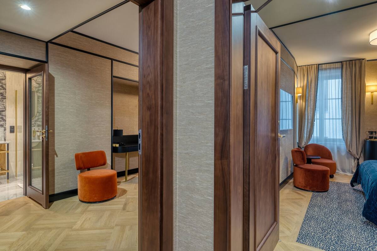 A modern hotel room with a wooden wardrobe, a small orange chair, a bed with a blue bedspread, and a desk with a computer.