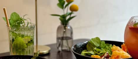 Una mesa de comedor con un bol de ensalada, un vaso de bebida y un pequeño florero con flores amarillas.