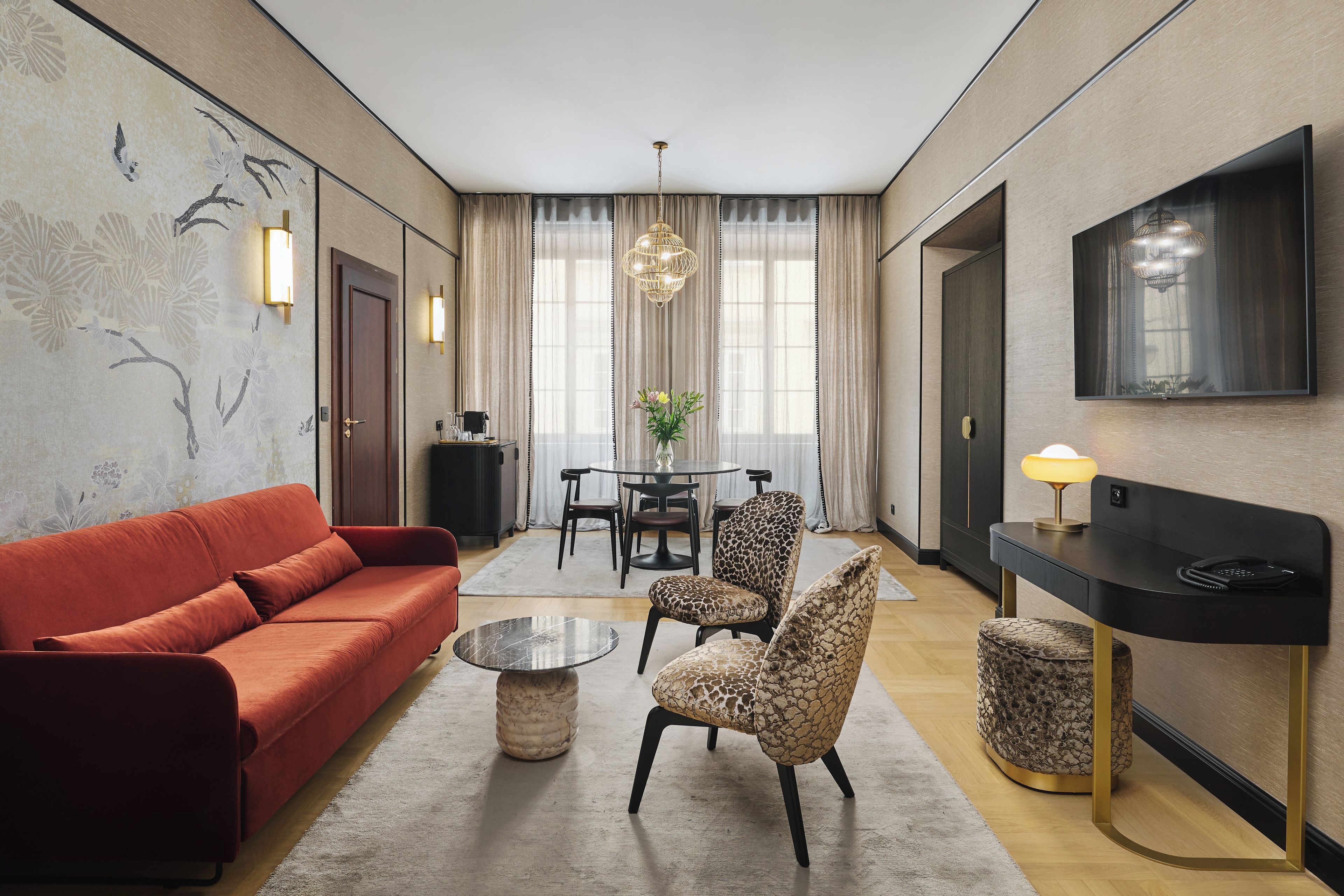 Un salon moderne avec un canapé rouge, une table basse ronde et des fauteuils à motifs. On y trouve également un coin repas avec une table et des chaises, ainsi qu’un téléviseur fixé au mur.