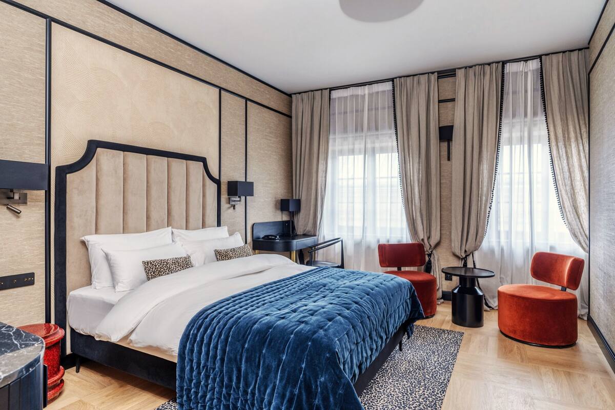 A modern hotel room with a large bed, a desk, two red chairs, and a large window with curtains.