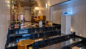 Un vestíbulo con escaleras de mármol, un arco dorado, un sillón amarillo y una columna blanca con un adorno azul.