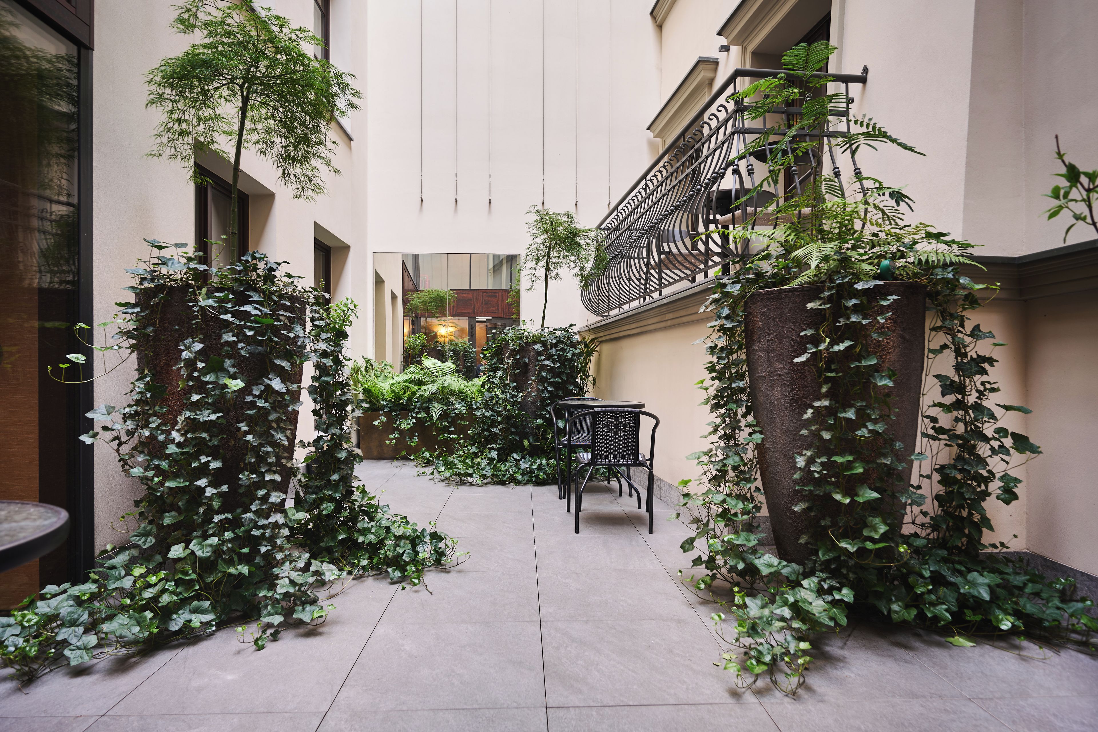 A narrow courtyard with a tiled floor, surrounded by walls with climbing plants. There is a small table and chairs in the center.