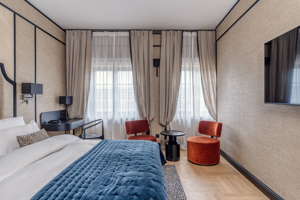 Una habitación de hotel moderna con una cama grande, dos sillas rojas, una mesita y un televisor montado en la pared.