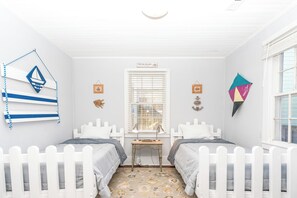 A room with two white beds, a small table, a window with blinds, and nautical-themed decorations.