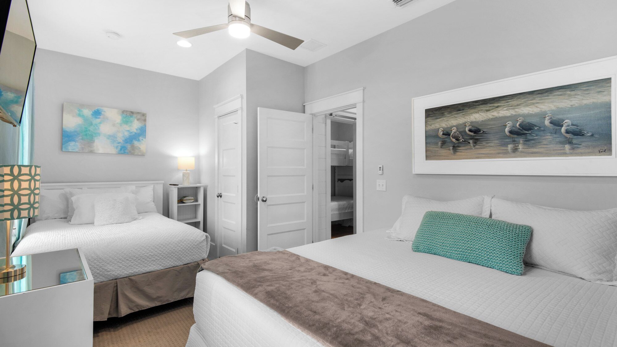 Habitación de hotel con dos camas, un ventilador de techo, un cuadro de gaviotas y una puerta que da a un armario.