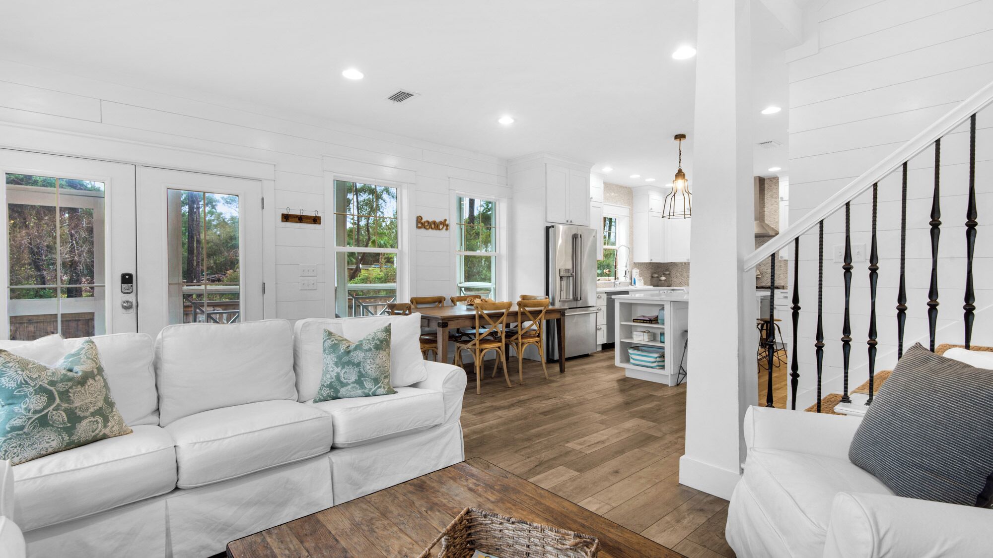A spacious living area with a white sofa, a dining table with chairs, a kitchen area with a refrigerator, and a staircase with black balusters.
