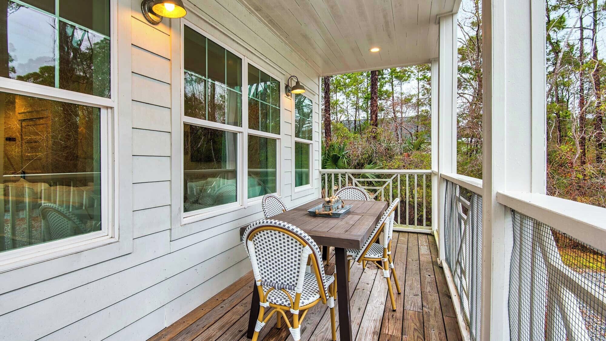 Un porche cubierto con una mesa y sillas de madera, ventanas grandes y vistas a árboles.