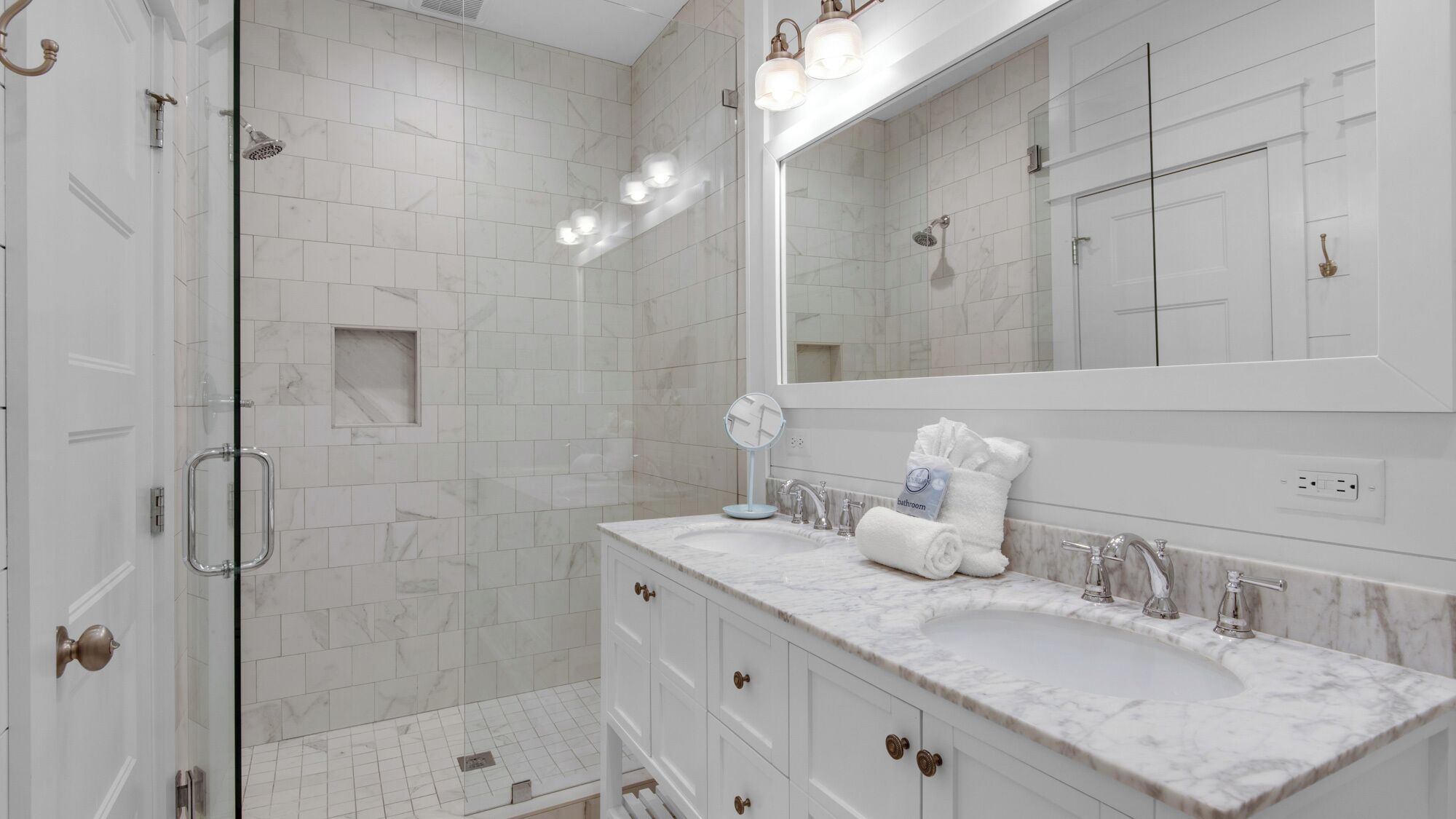 A modern bathroom with white marble tiles, a large mirror, a glass shower enclosure, and a vanity with a sink and towel rack.