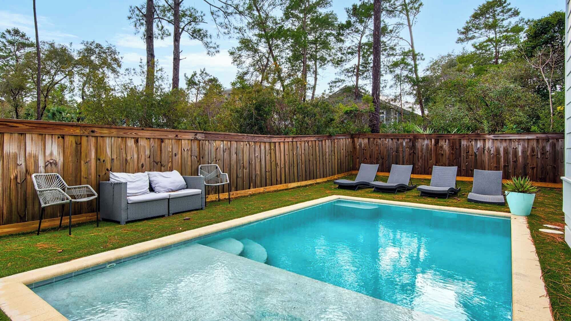 Ein Garten mit Swimmingpool, Holzzaun, grauem Sofa und Liegestühlen.