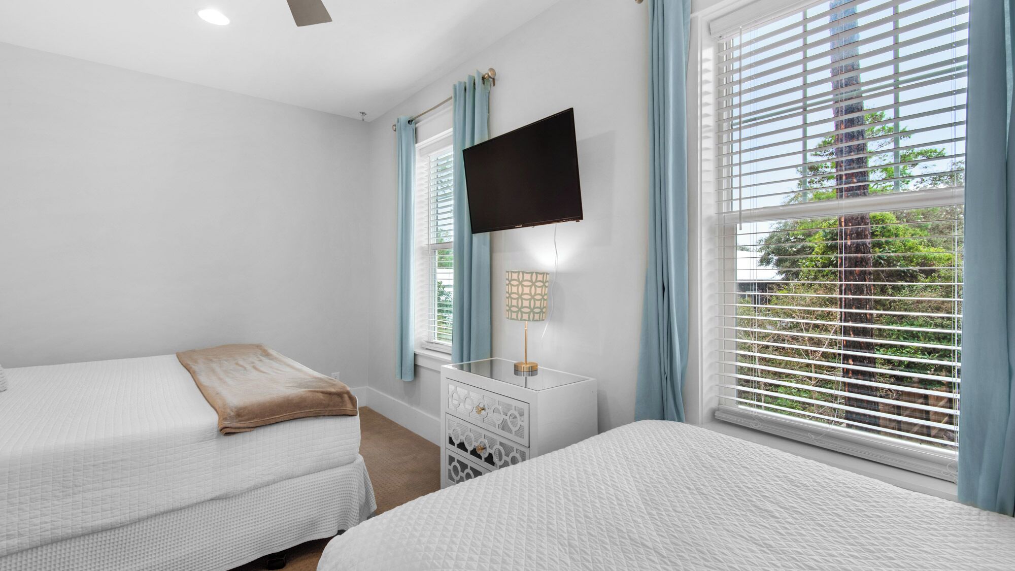 A bedroom with two beds, a TV mounted on the wall, a bedside table with a lamp, and large windows with blinds.