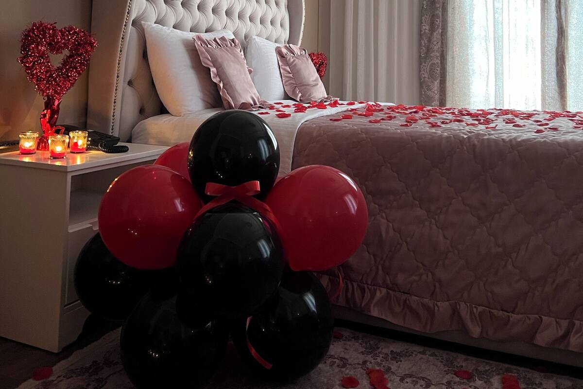 Ein Schlafzimmer mit einer gepolsterten Kopfteile, einem Nachttisch mit Kerzen und einem Bett mit Rosenblättern und Luftballons.
