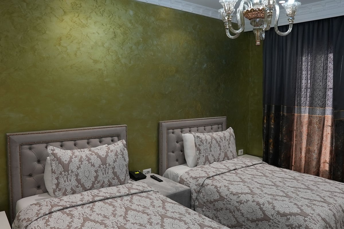 Ein Hotelzimmer mit zwei Betten, einer Kristallleuchter und Blick durch Vorhänge nach draußen.