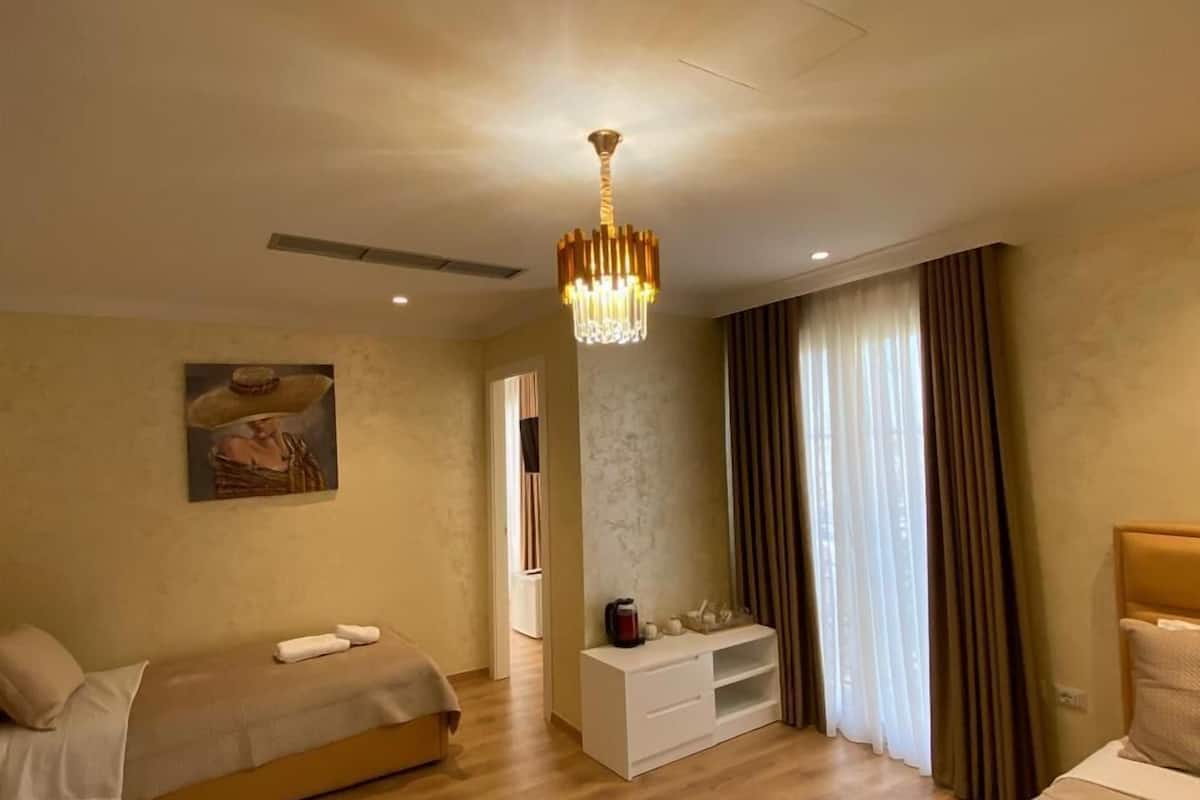 Ein Hotelzimmer mit Doppelbett, Einzelbett, Schminktisch mit Spiegel, einer Chandelüster und einem Gemälde an der Wand.