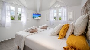 Ett sovrum med en säng, två fönster med fönsterluckor, en tv på väggen och utsikt över havet.