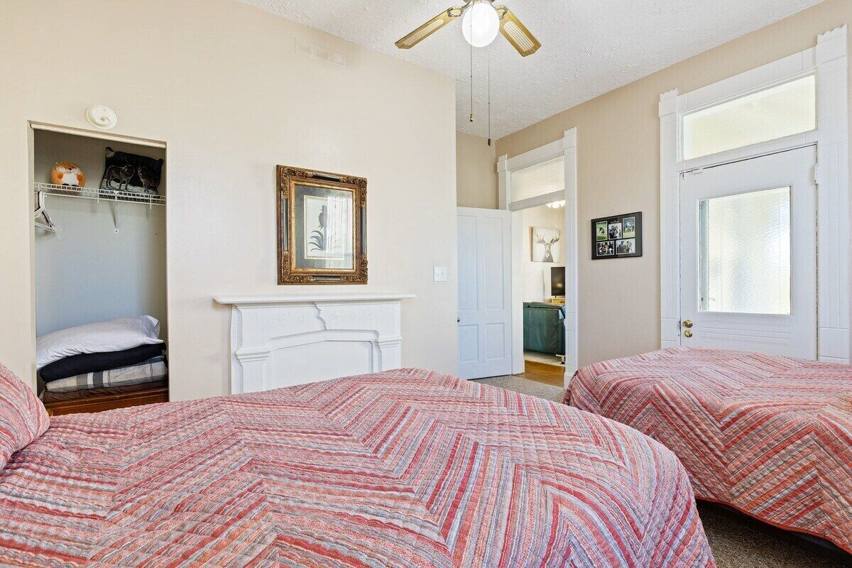 A bedroom with two beds, a white fireplace, a ceiling fan, and a door leading to a bathroom.