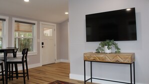 Un salon moderne avec un téléviseur à écran plat fixé au mur, une table basse en bois agrémentée d’une plante, et un coin repas avec des chaises et une table.