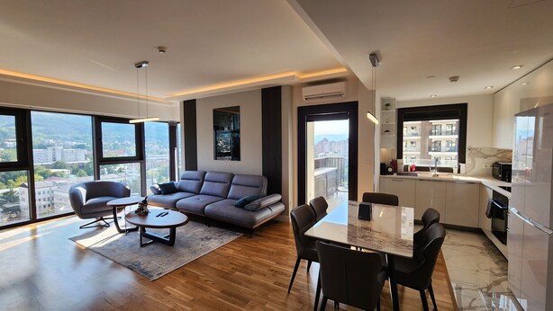 A modern living room with a sofa, chairs, a dining table, and a kitchen area with a view of the city. - Diamond Residences 2bd 2bth 11th fl. Lux Apt w/ Free Parking (Skopje)