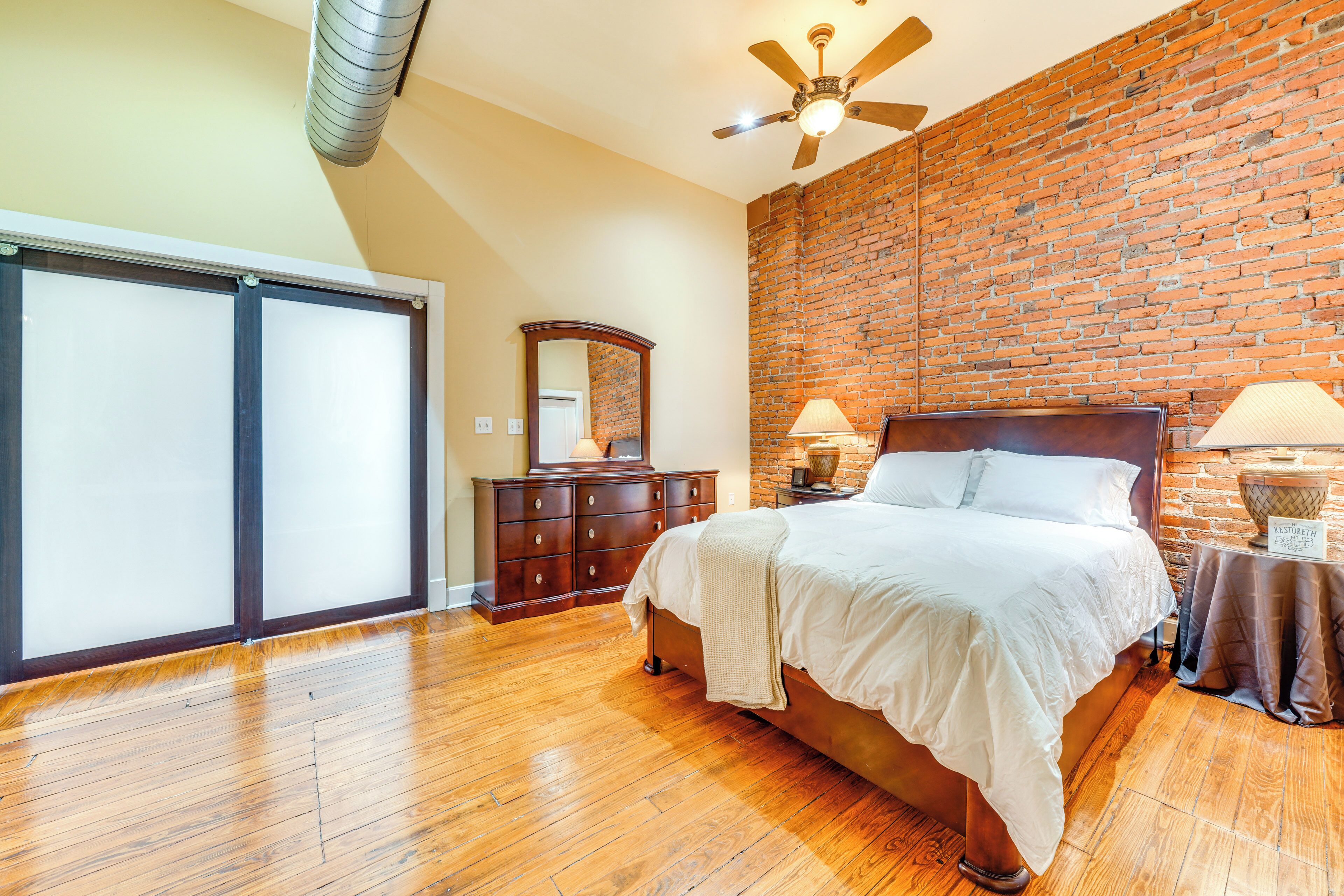 Quarto com parede de tijolos, piso de madeira, cama, cômoda e ventilador de teto.