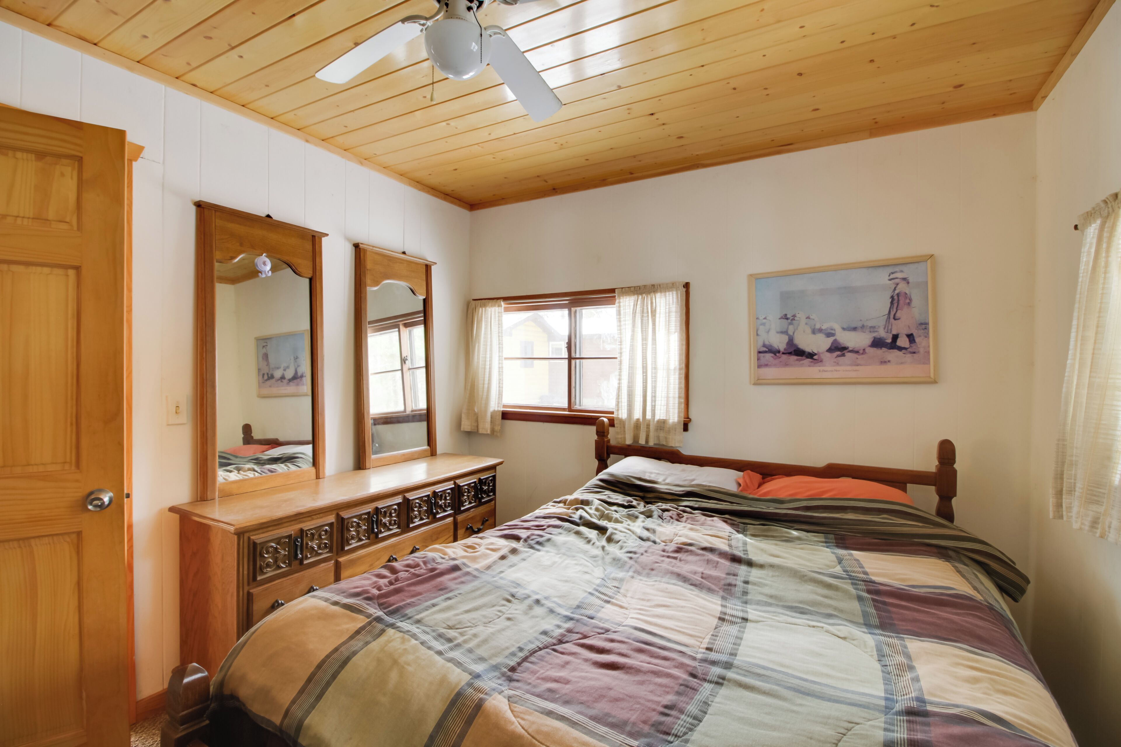 A bedroom with a wooden ceiling, a bed with a plaid bedspread, a dresser with drawers, a mirror, and a window with curtains.