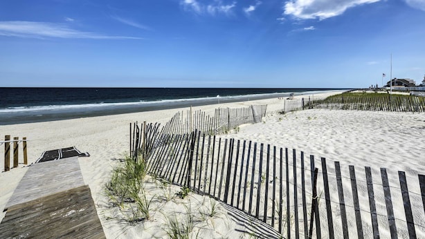 Une plage avec une promenade en bois menant à l’eau, une zone sablonne délimitée par une clôture et un bâtiment en arrière-plan.