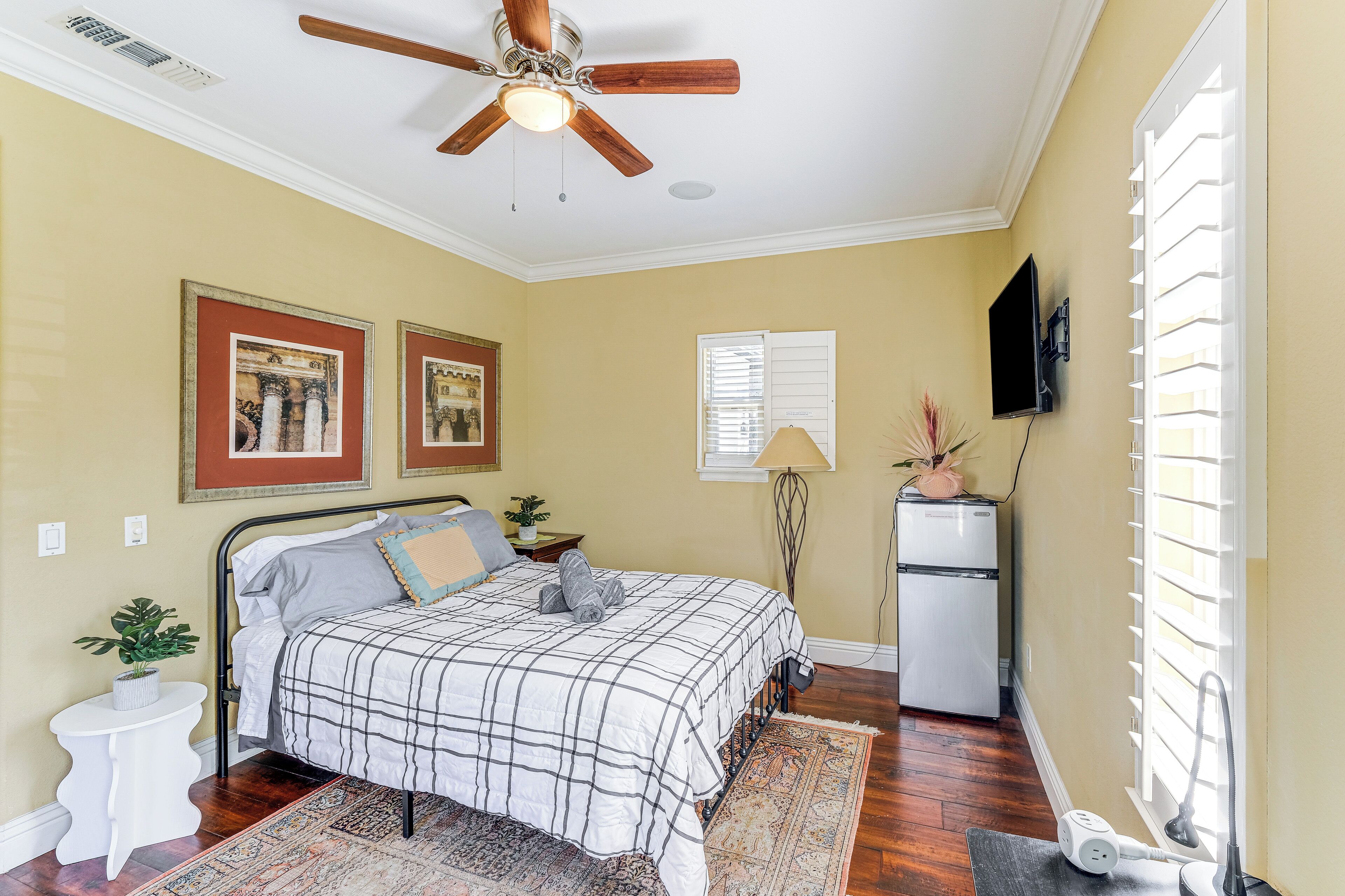 Une chambre à coucher comprenant un lit, un ventilateur de plafond, une télévision, un réfrigérateur et une fenêtre avec des stores.