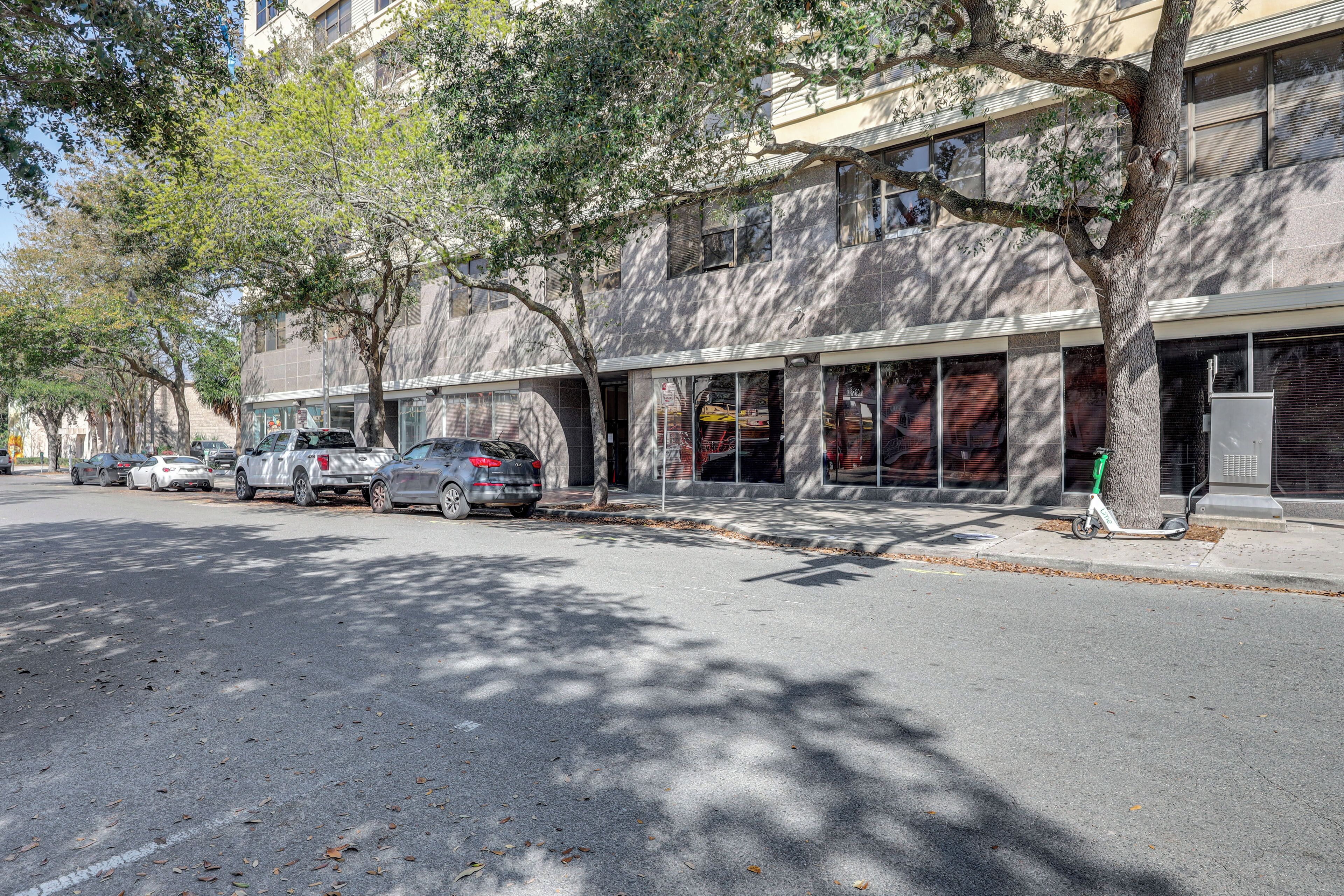A street view with parked cars, trees, and a building with large windows and a storefront.