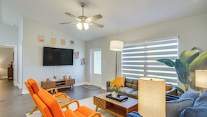A spacious living room with a ceiling fan, a flat-screen TV mounted on the wall, a sofa with a lamp, and a dining area with orange chairs. - 7 Mi to River District: Cape Coral Home w/ Lanai (Cape Coral)